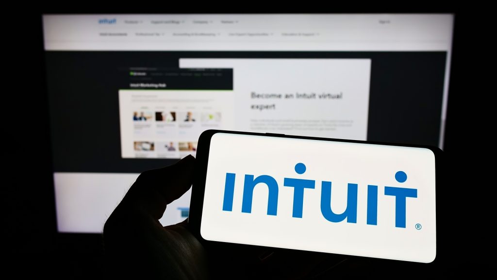 A logo of Intuit on a screen of a horizontally flipped smartphone; at the back is a computer screen showing Intuit’s website homepage.